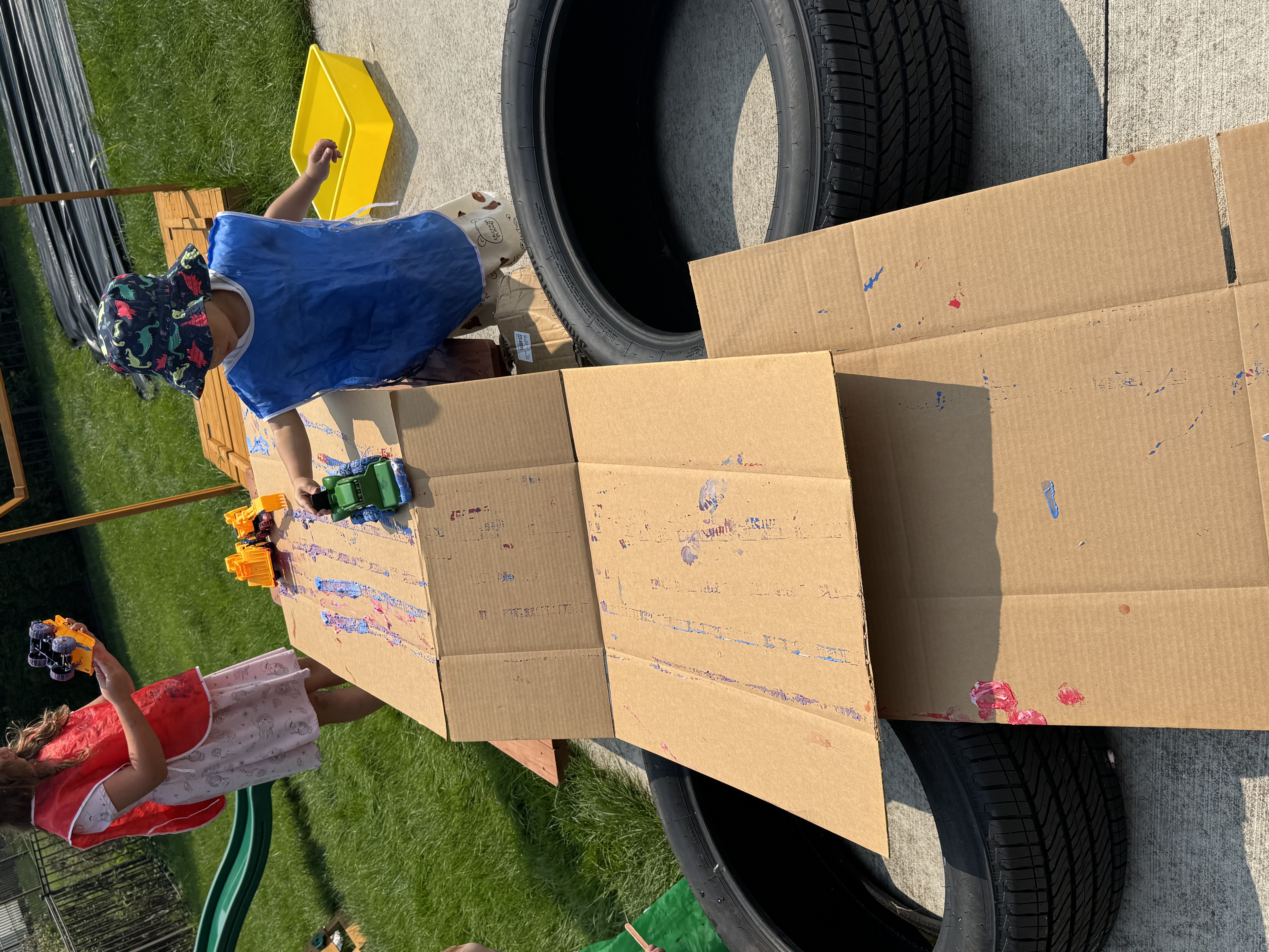 Children playing with paint, toy cars, and cardboard outside