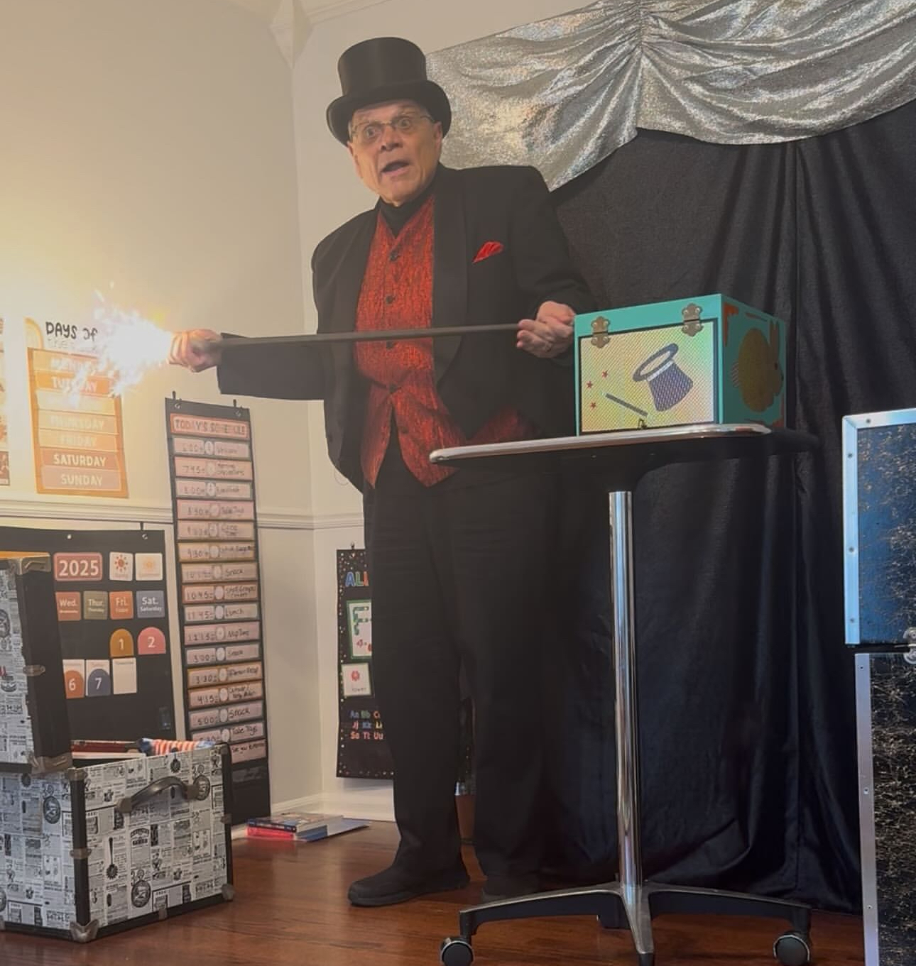 An old man in a magician costume, holding a wand, standing next to a box on a table