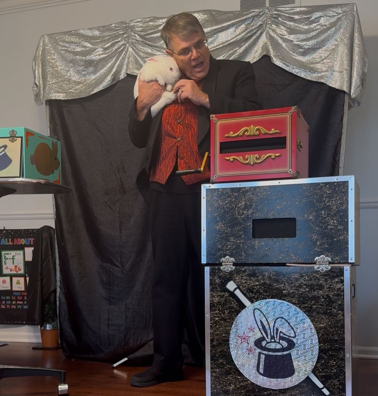 An old man in a magician costume holding a white bunny, standing next to boxes